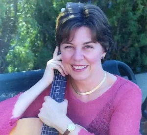 Josee Vachon smiles while holding her guitar in this headshot. Josee has short brown hair and is wearing a pink shirt.